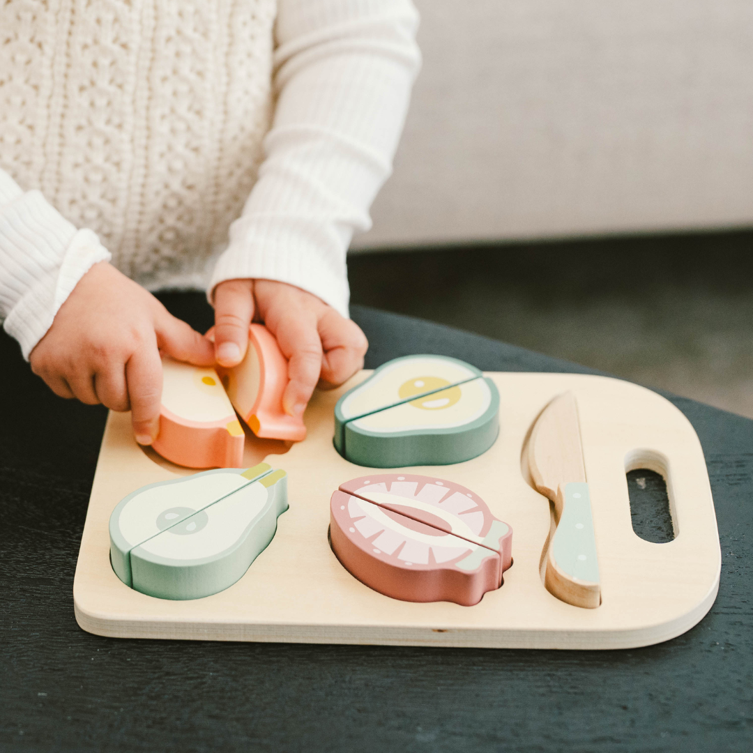 Girl´s hands playing with a wooden cutting board with colorful, realistic-looking fruit pieces arranged on top of it, and a wooden knife beside them.