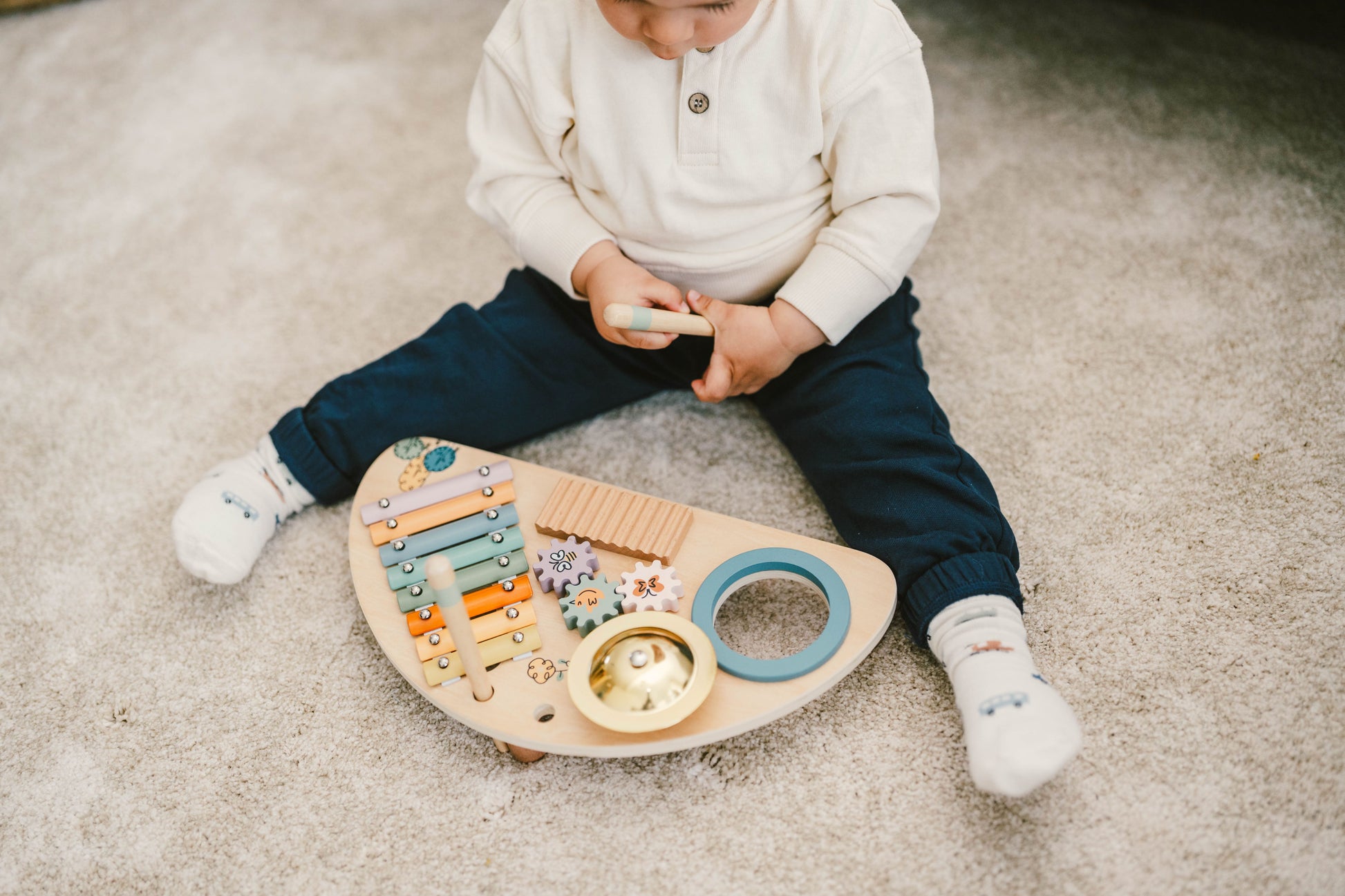 Boy playing with a wooden music center toy with multiple colorful instruments and interactive elements, including a xylophone, cymbal, guiro, and spinning gears, along with drumsticks. The toy is crafted from natural wood with pastel colors and nature-themed illustrations.