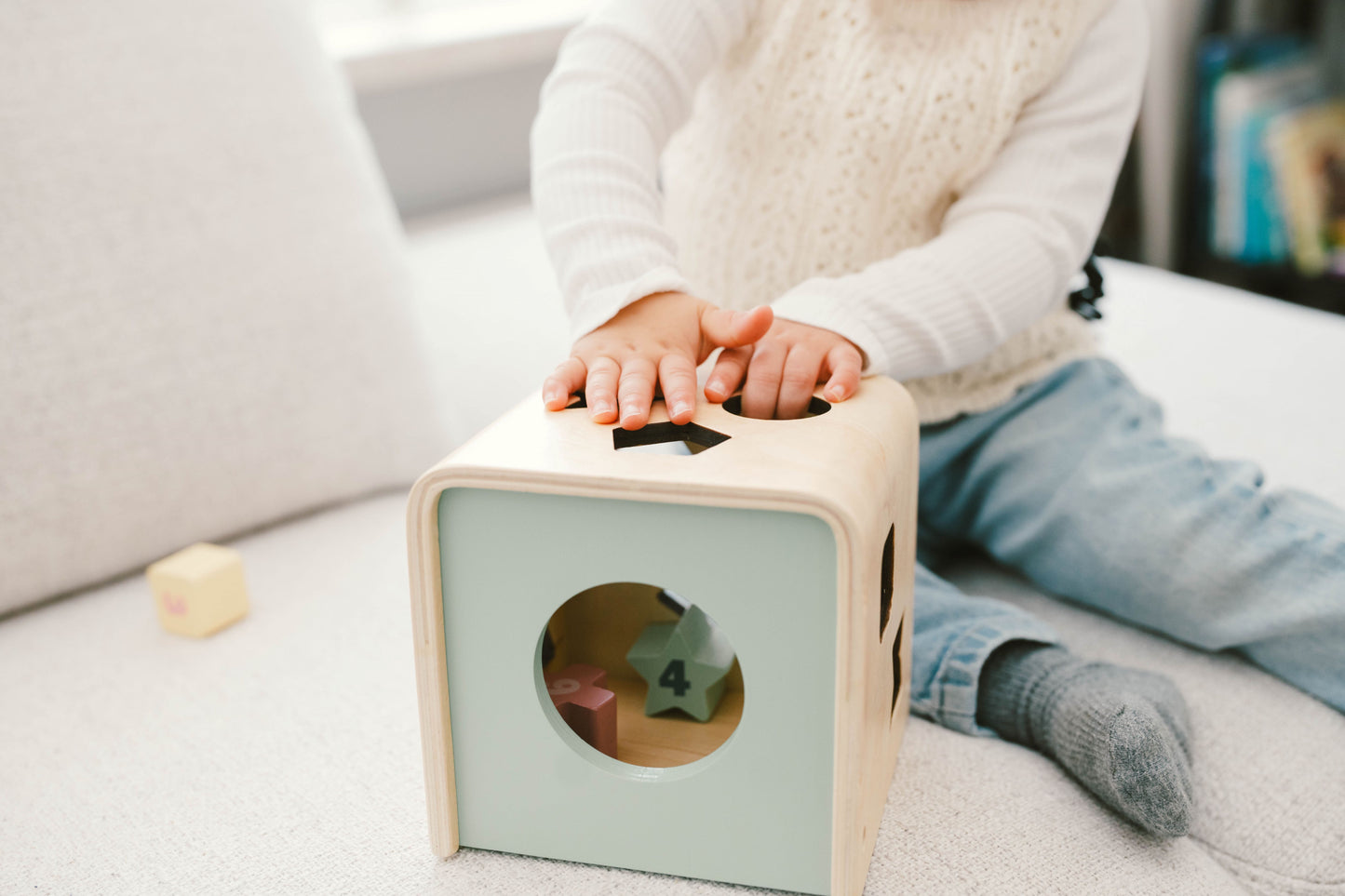 A wooden shape sorting cube with various holes for shapes, and colorful numbered shapes being inserted by a hand.