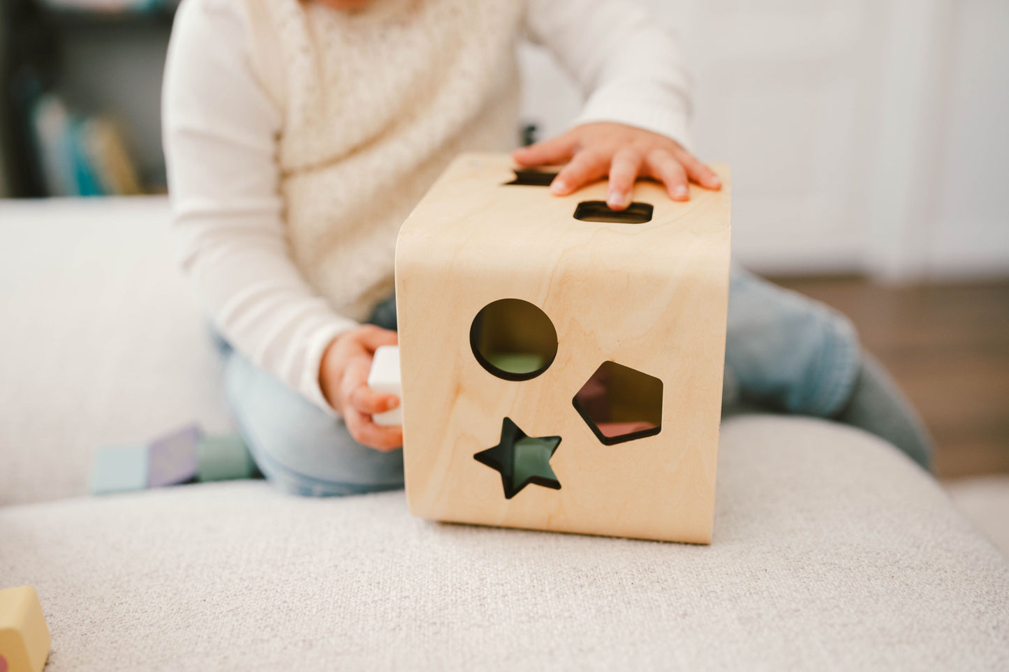 A wooden shape sorting cube with various holes for shapes, and colorful numbered shapes being inserted by a hand.