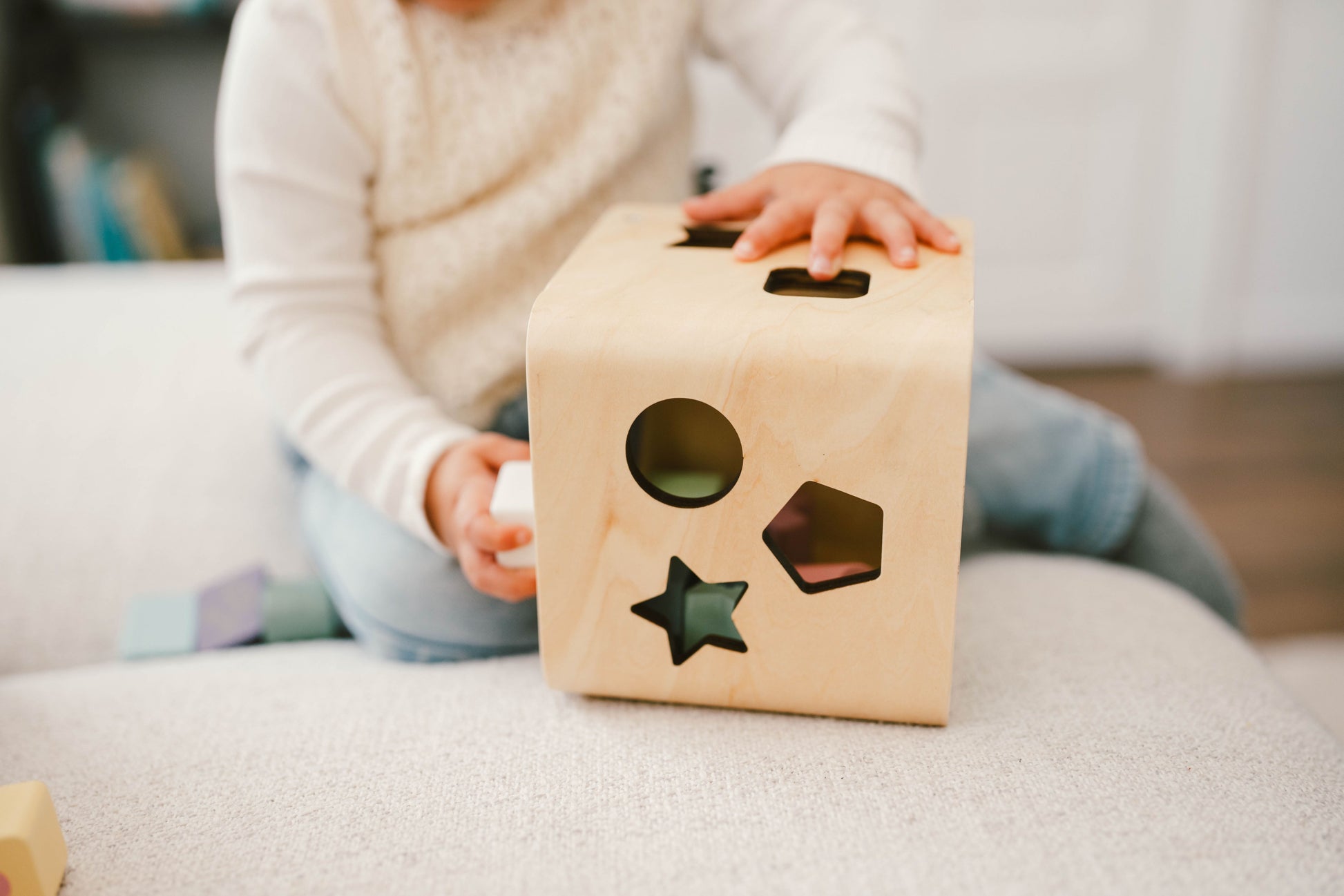 A wooden shape sorting cube with various holes for shapes, and colorful numbered shapes being inserted by a hand.