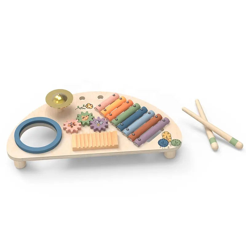 A wooden music center toy with multiple colorful instruments and interactive elements, including a xylophone, cymbal, guiro, and spinning gears, along with drumsticks. The toy is crafted from natural wood with pastel colors and nature-themed illustrations.
