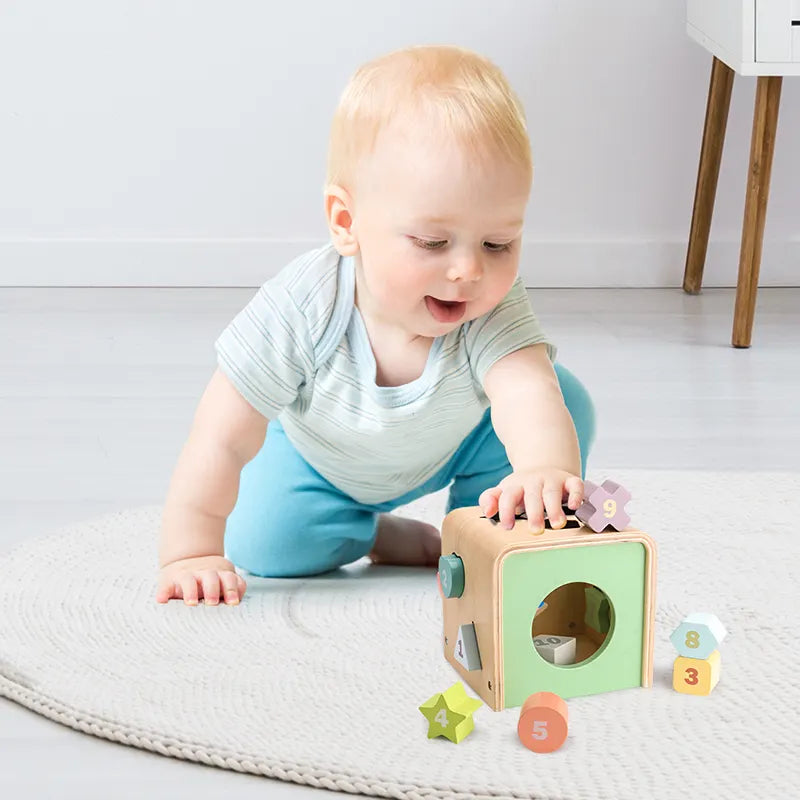 A wooden shape sorting cube with various holes for shapes, and colorful numbered shapes being inserted by a hand.